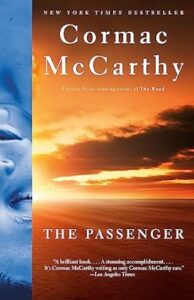The image shows the cover of a book titled "the passenger" by author cormac mccarthy, recognized as a new york times bestseller and penned by the pulitzer prize-winning author of "the road." the cover features a merging of two contrasting images: a detailed, up-close picture of a person's face on the left side, and a scenic sunset over a desolate landscape on the right side. prominently displayed at the bottom is a review from the los angeles times, praising the book with the words "a brilliant book... as stunning as only cormac mccarthy can.