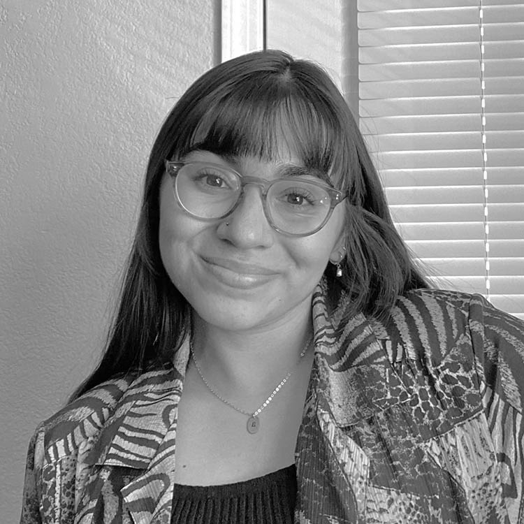 A monochromatic portrait of a smiling woman wearing glasses, a patterned shirt, and a delicate necklace, sitting indoors with blinds in the background.