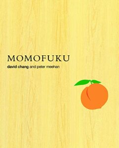 A book cover with a wooden background texture featuring the title "momofuku" in bold black letters at the top, with the authors' names "david chang and peter meehan" just below, and an illustration of an orange peach with two green leaves towards the bottom right.