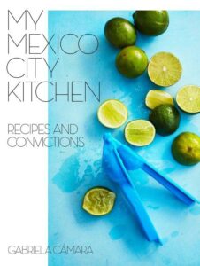 A colorful assortment of fresh limes and a blue juicer on a light blue background, symbolizing the vibrant flavors of mexican cuisine, as featured on the cover of "my mexico city kitchen: recipes and convictions" by gabriela cámara.
