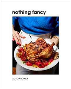 A person in a blue top presenting a plate with a roasted chicken and tomatoes, with the title "nothing fancy" displayed above.