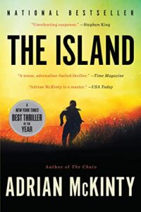 A book cover for "the island" by adrian mckinty, featuring praise from stephen king and time magazine, and noted as a new york times best thriller of the year, set against a backdrop of a silhouette of a person in a field at sunset.