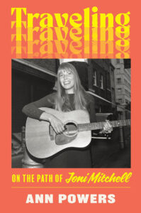 A book cover titled "traveling on the path of joni mitchell" by ann powers, featuring a black and white photo of joni mitchell smiling and playing a guitar on a city street. vibrant red and yellow text overlays the image.