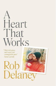 Cover of the book "A Heart That Works" by Rob Delaney. The title and author’s name are displayed prominently. Below the title is a photograph of a man and young child embracing outside in a snowy setting. A quote by Anne Lamott is also visible.