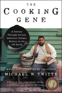 Cover of "The Cooking Gene" by Michael W. Twitty, featuring the author seated with a backdrop of an old wooden building.