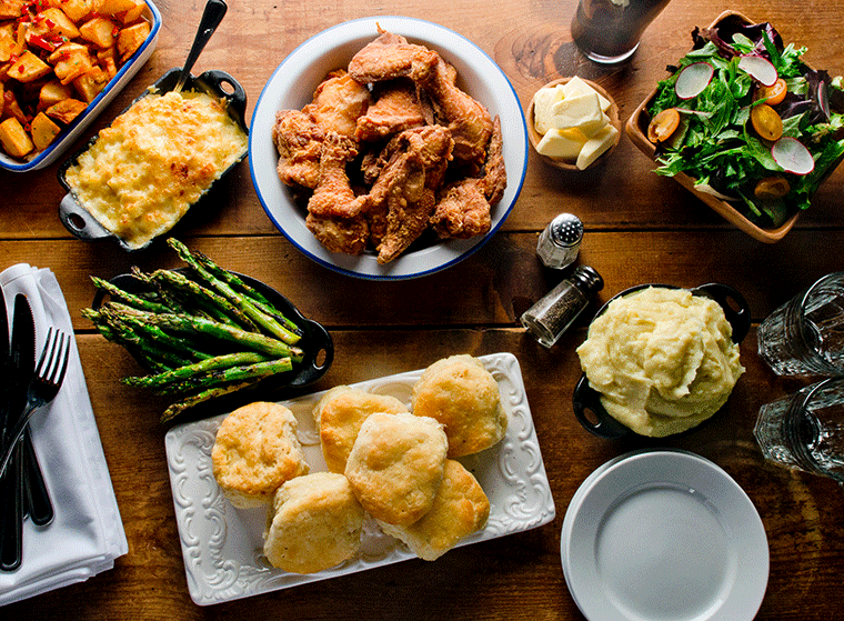 A table with fried chicken, mashed potatoes, biscuits, asparagus, salad, roasted potatoes, and mac and cheese.