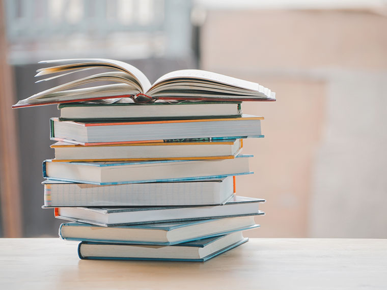 A stack of books with an open book on top, placed on a wooden surface.