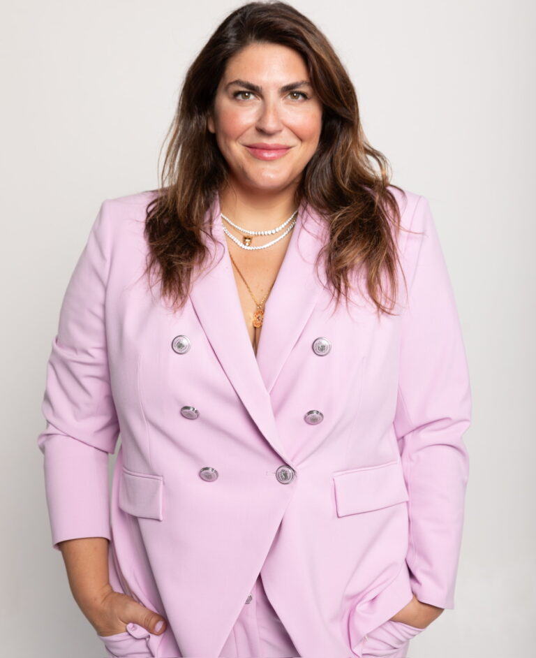 Person in a light pink suit, smiling with hands in pockets against a plain background.