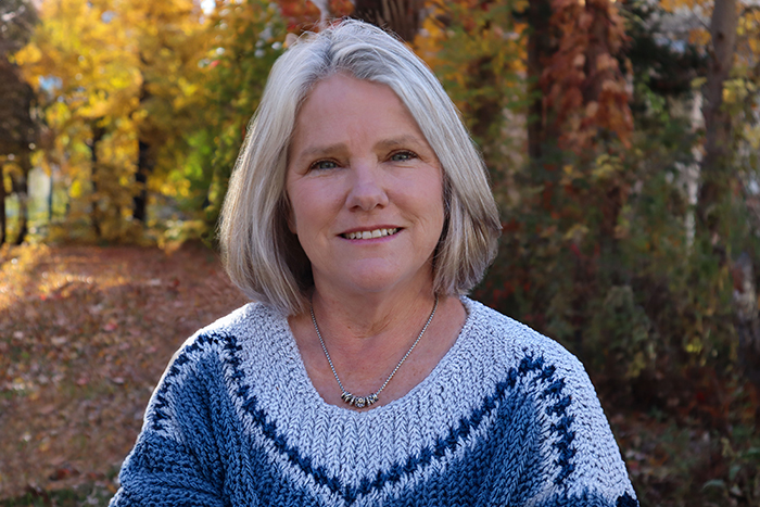 Smiling woman with gray hair in a blue and white sweater, set against an autumnal background.