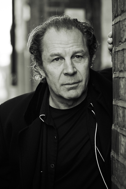 Black and white portrait of an older man with wavy hair, leaning against a brick wall, looking serious.
