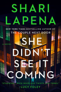 Book cover for "She Didn't See It Coming" by Shari Lapena with lit apartment windows and silhouettes of people inside.