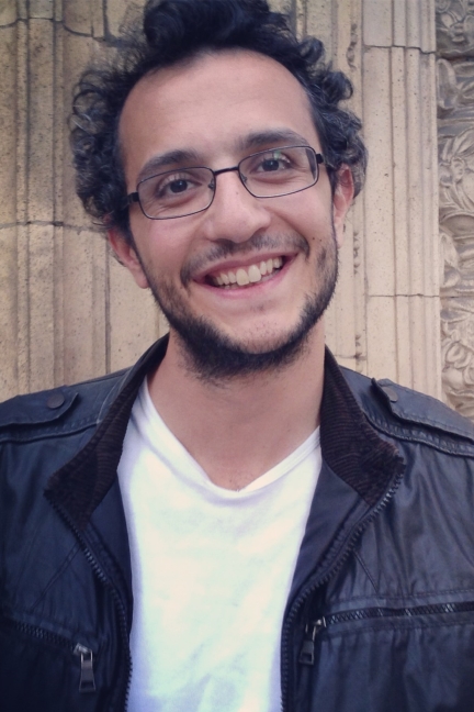 A smiling man with glasses and curly hair wears a black jacket and white shirt, standing in front of a stone wall.