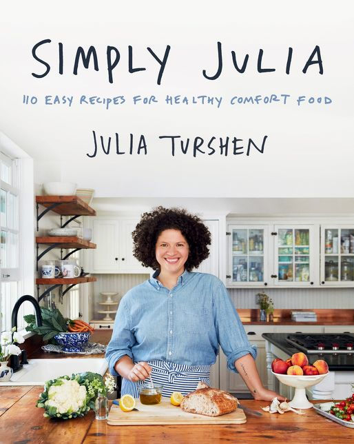 A smiling woman stands in a bright kitchen with fresh produce and bread on the counter in front of her.