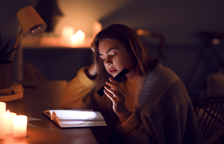 Woman reading a book by candlelight, using a flashlight, wearing a sweater, sitting at a table at night.
