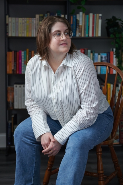A person in glasses sits on a chair, wearing a striped shirt and jeans, with bookshelves in the background.