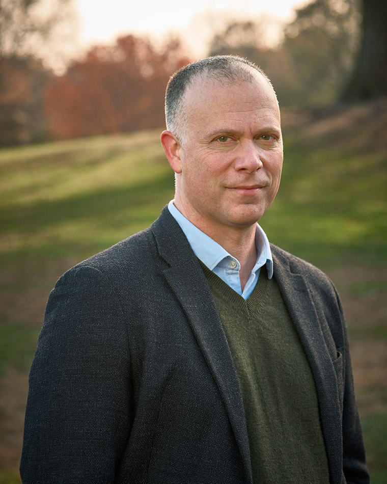 A man in a blazer stands outside on a grassy field with trees in the background at sunset.
