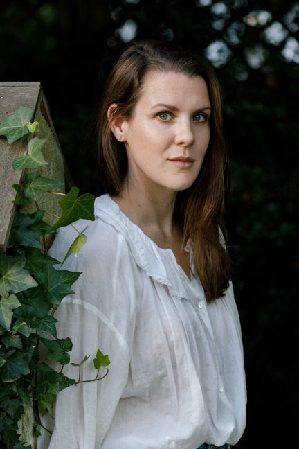 Woman with long brown hair in a white blouse stands by a wooden fence with green ivy, outdoors.