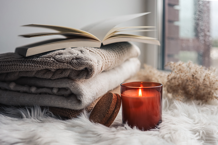 A lit candle, folded sweaters, and an open book rest on a soft fuzzy surface by a window.