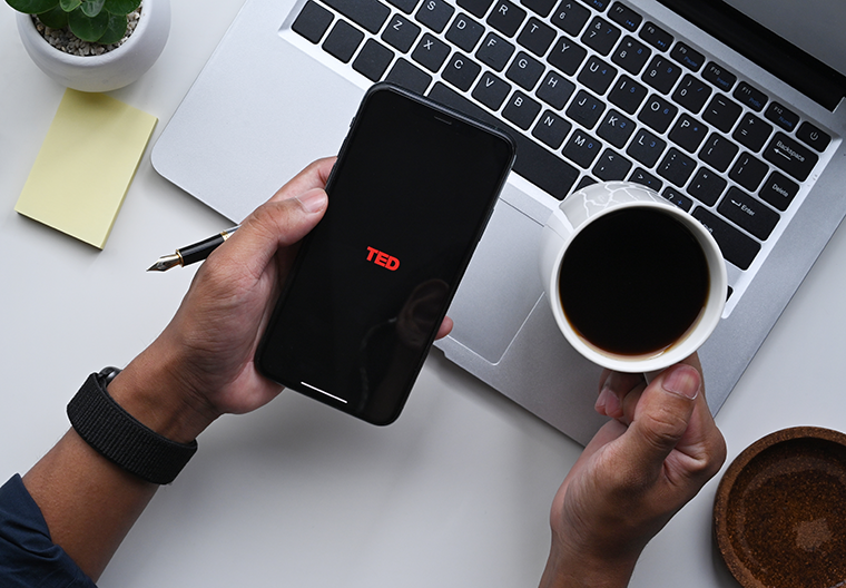 Person holding a phone with the TED logo and a cup of coffee near a laptop on a white desk.