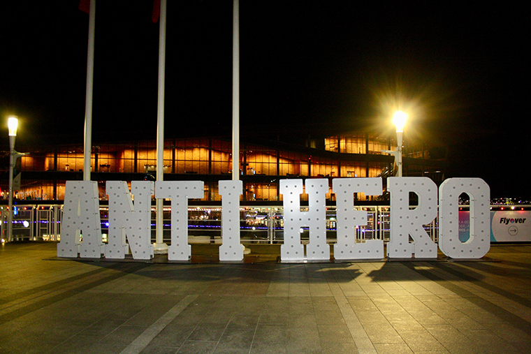 Large illuminated letters spelling "ANTI HERO" displayed outdoors at night with a lit building in the background.