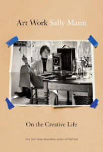 Book cover of "Art Work" by Sally Mann, featuring a taped photo of a woman with a vintage camera and desk items.