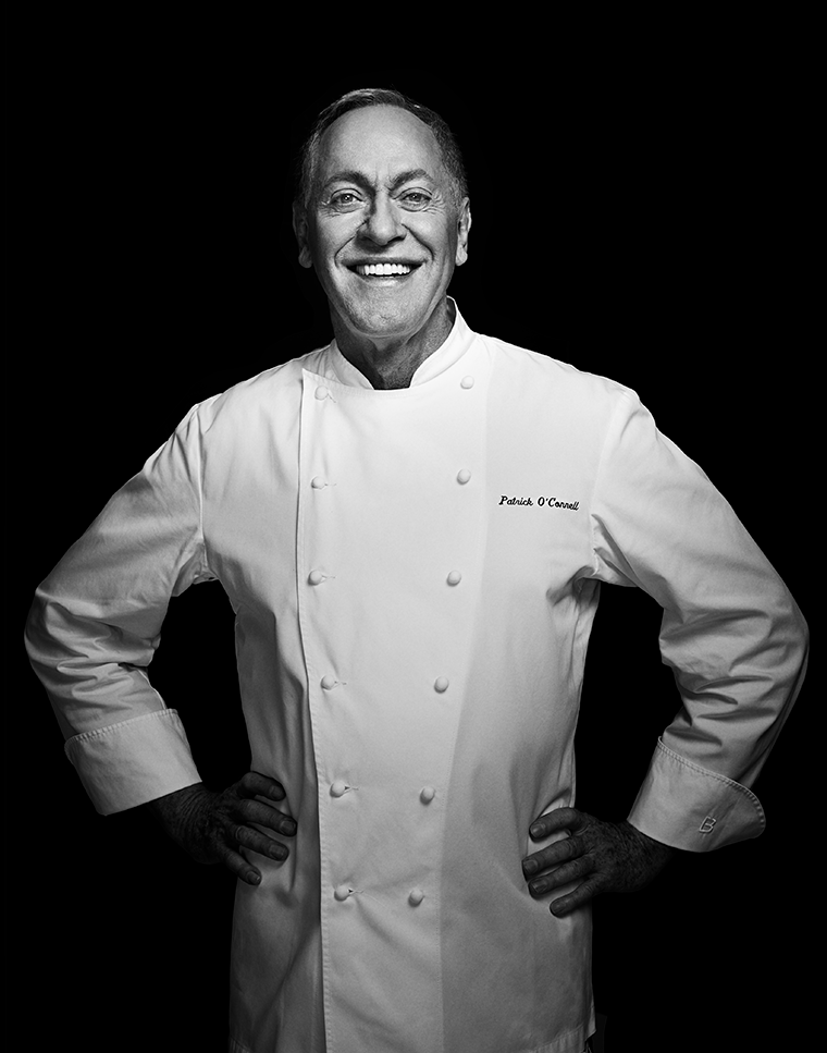 A smiling chef in a white uniform stands with hands on hips against a black background.