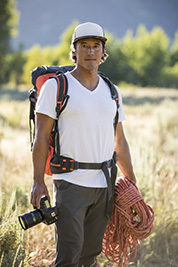 Man outdoors in hiking gear holding a camera and climbing rope, standing in a sunlit field with trees behind.