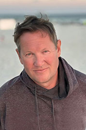 Man with short brown hair wearing a dark hoodie standing on a beach with a calm expression.