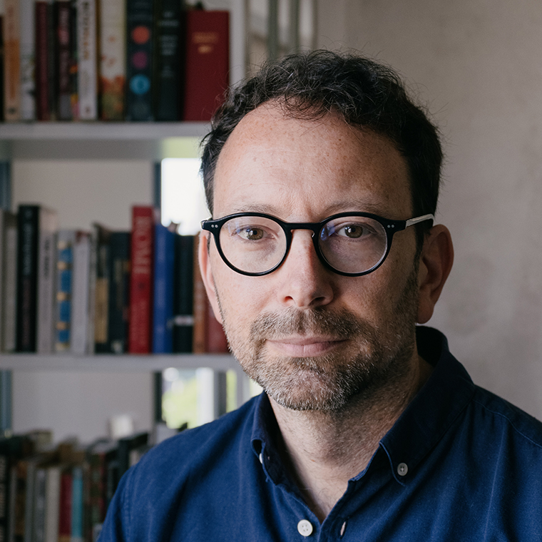 Man with glasses and beard in blue shirt, sitting in front of a bookshelf filled with books.