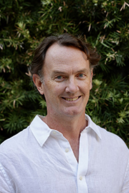 Man in a white shirt smiling in front of green foliage.