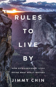 A person stands on a cliff edge at sunset with mountains in the background; text reads "Rules to Live By" by Jimmy Chin.
