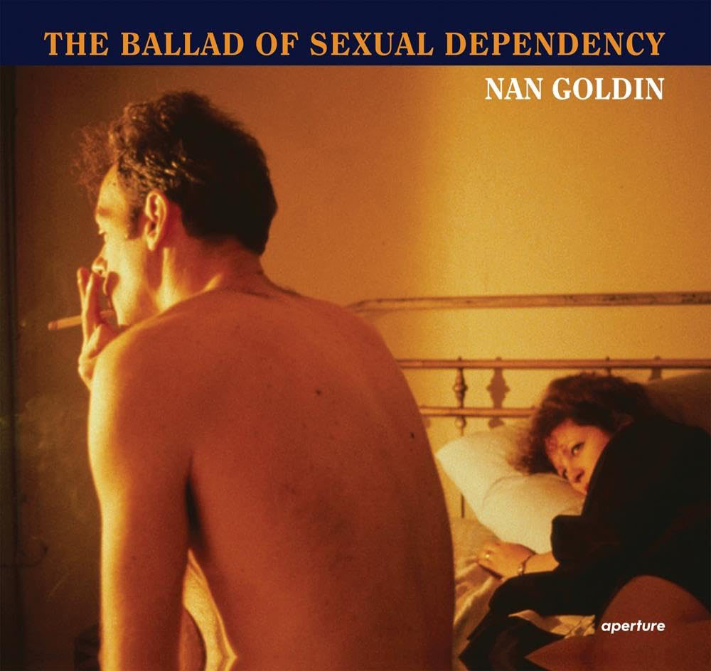 A shirtless man smokes on a bed while a woman lies beside him, gazing toward the camera.