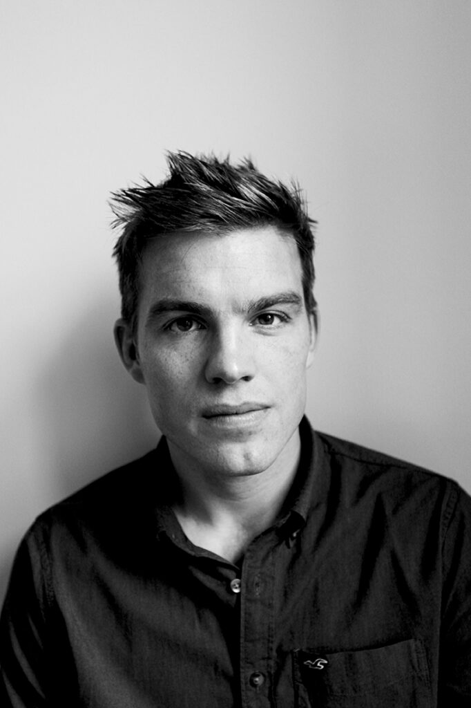 Black and white portrait of a young man with short spiked hair, wearing a dark button-up shirt.