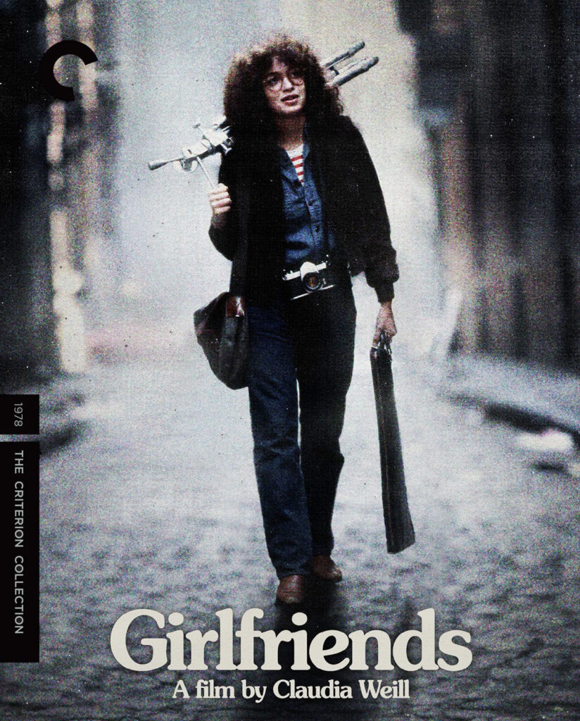 A woman with curly hair walks down a street carrying a tripod and camera case; "Girlfriends" is the film title.
