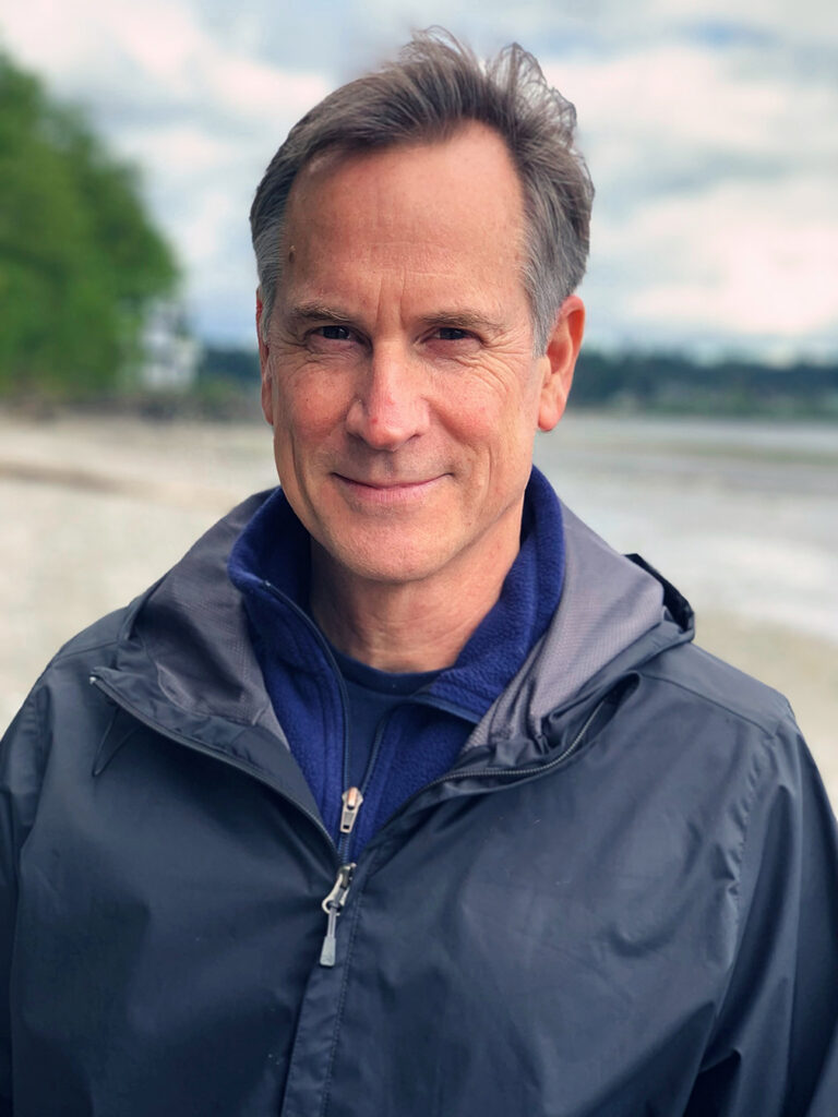 Man in a dark jacket smiles outdoors near a beach, with trees and water in the background.