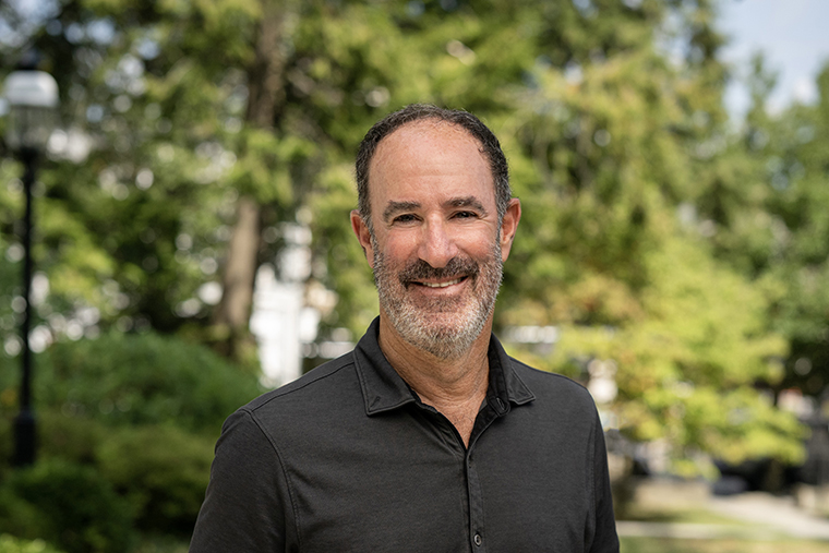 Smiling man with short dark hair and beard, wearing a dark shirt, stands outside with trees in background.